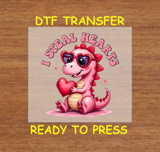 "I Steal Hearts Dinosaur" DTF Transfer featuring a cute pink dinosaur with heart sunglasses holding a red heart, perfect for Valentine’s Day crafting.